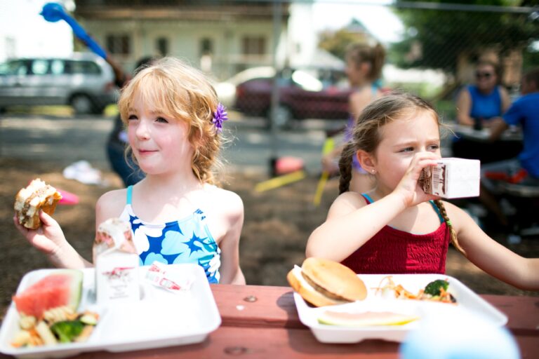 Two children sit at a picnic table, eating and drinking