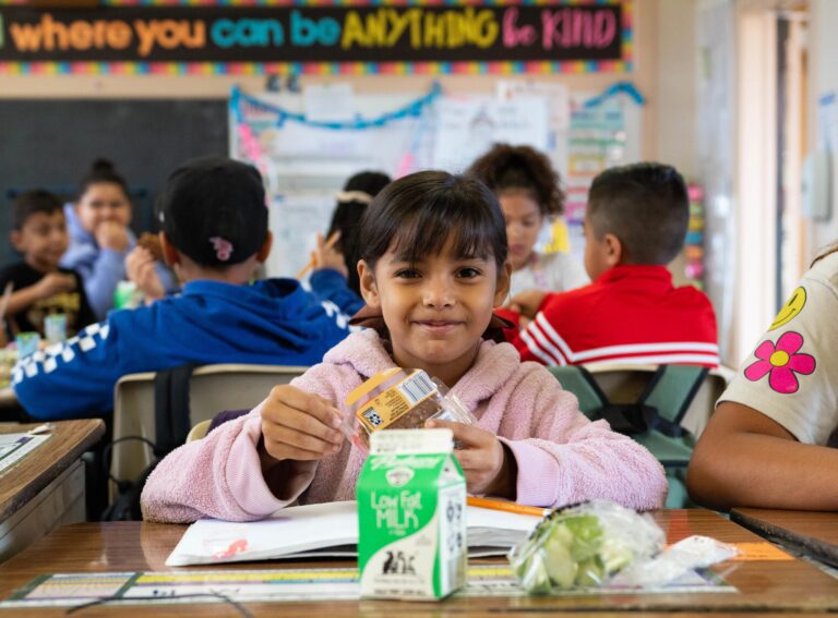 An elementary school girl sits at her desk and smiles, holding a breakfast item.