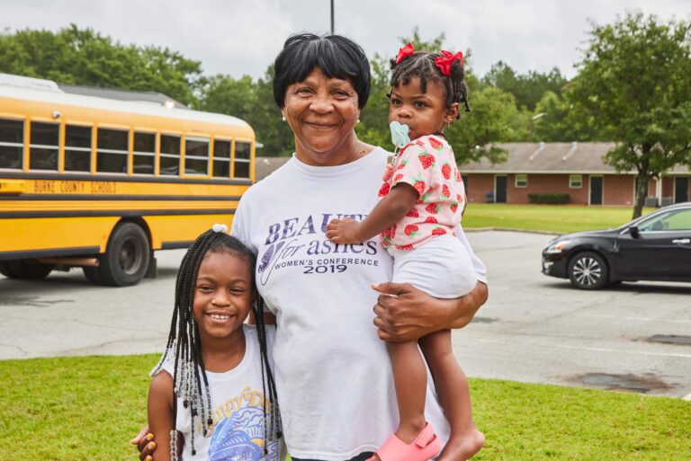 A caregiver holds one child and has her arm around another. They smile and stand in front of a school bus.