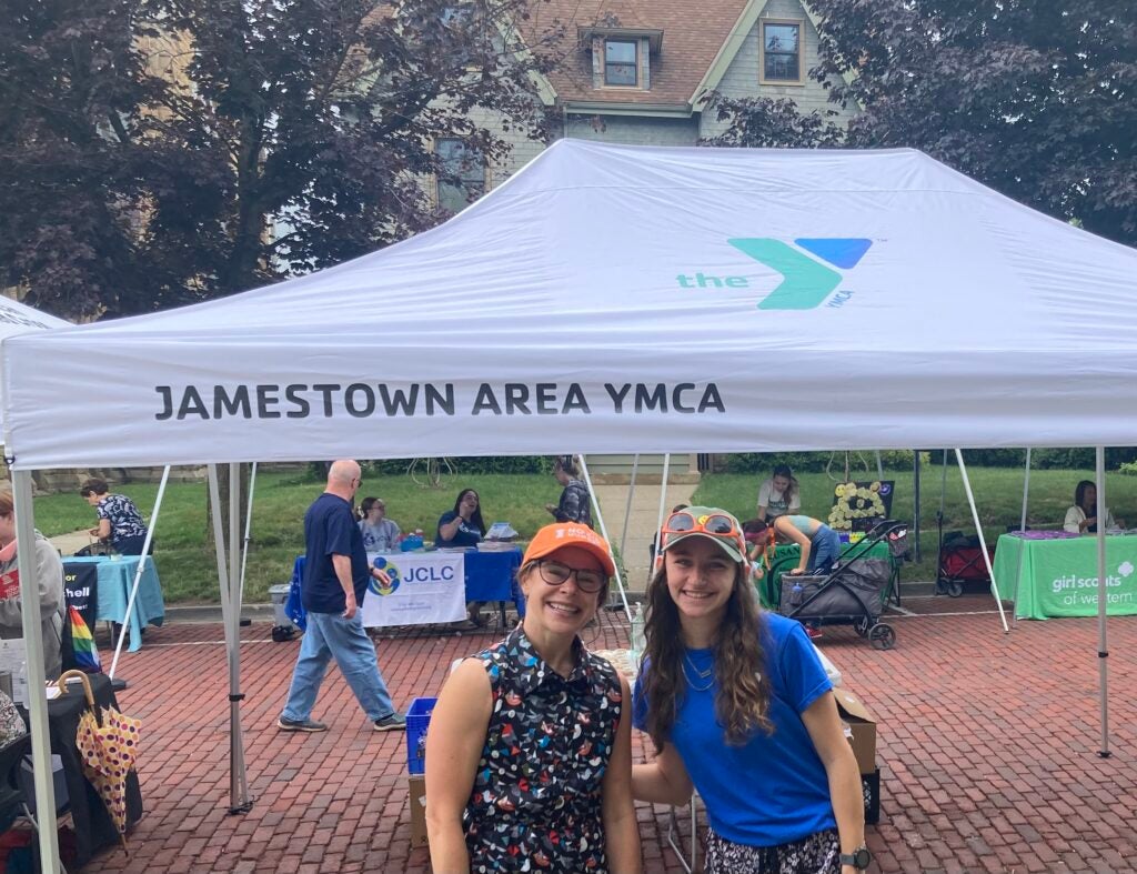 No Kid Hungry NY Program Manager Liz and Youth Ambassador Acacia smile in front of a pop up tent with the Jamestown Area YMCA logo