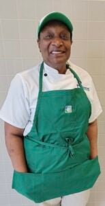 Lenor, with her hands in her apron pockets, smiles at the camera