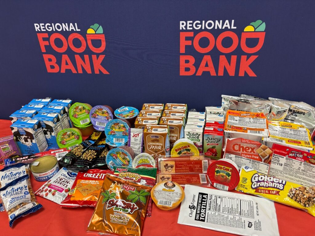 Background has the Regional Food Bank logo; in front, a table is covered with all sorts of food: milk, cereal, apple sauce, and more