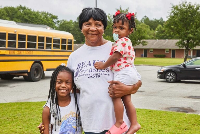 A caregiver holds one child with her arm around another, standing outside in front of a school bus