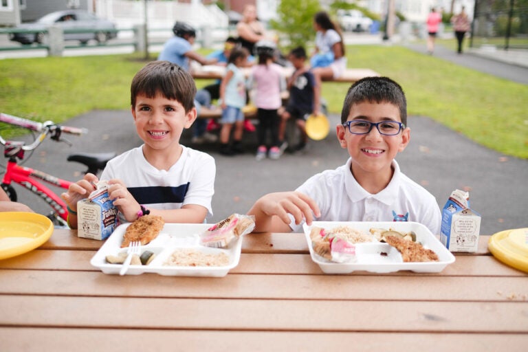 Two young children sit at a picnic table, smiling at the camera with trays of food in front of them